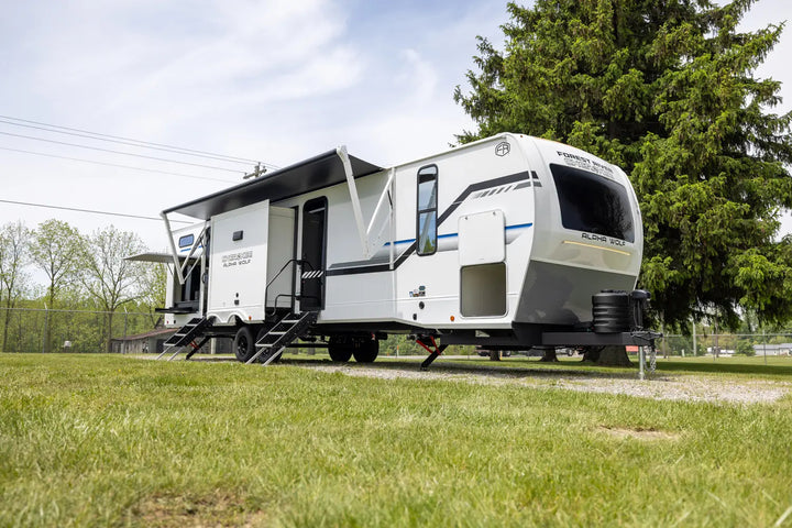 White travel trailer with an extended awning parked on grass with trees in the background